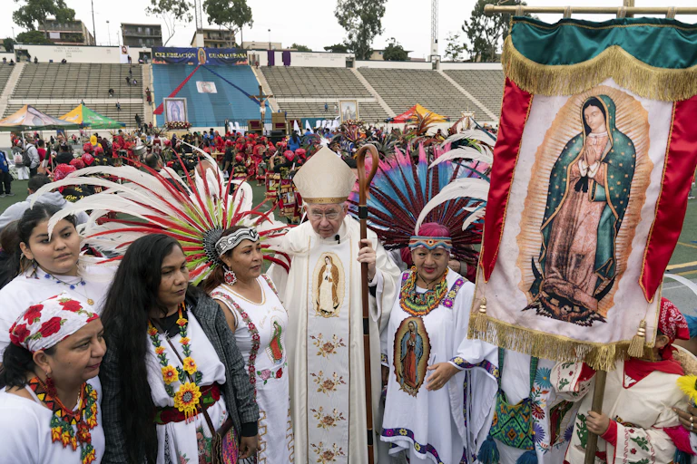 A man in white clerical vestments stands in an outdoor stadium, surrounded by women in white outfits holding a banner with a picture of a woman in a blue cloak.