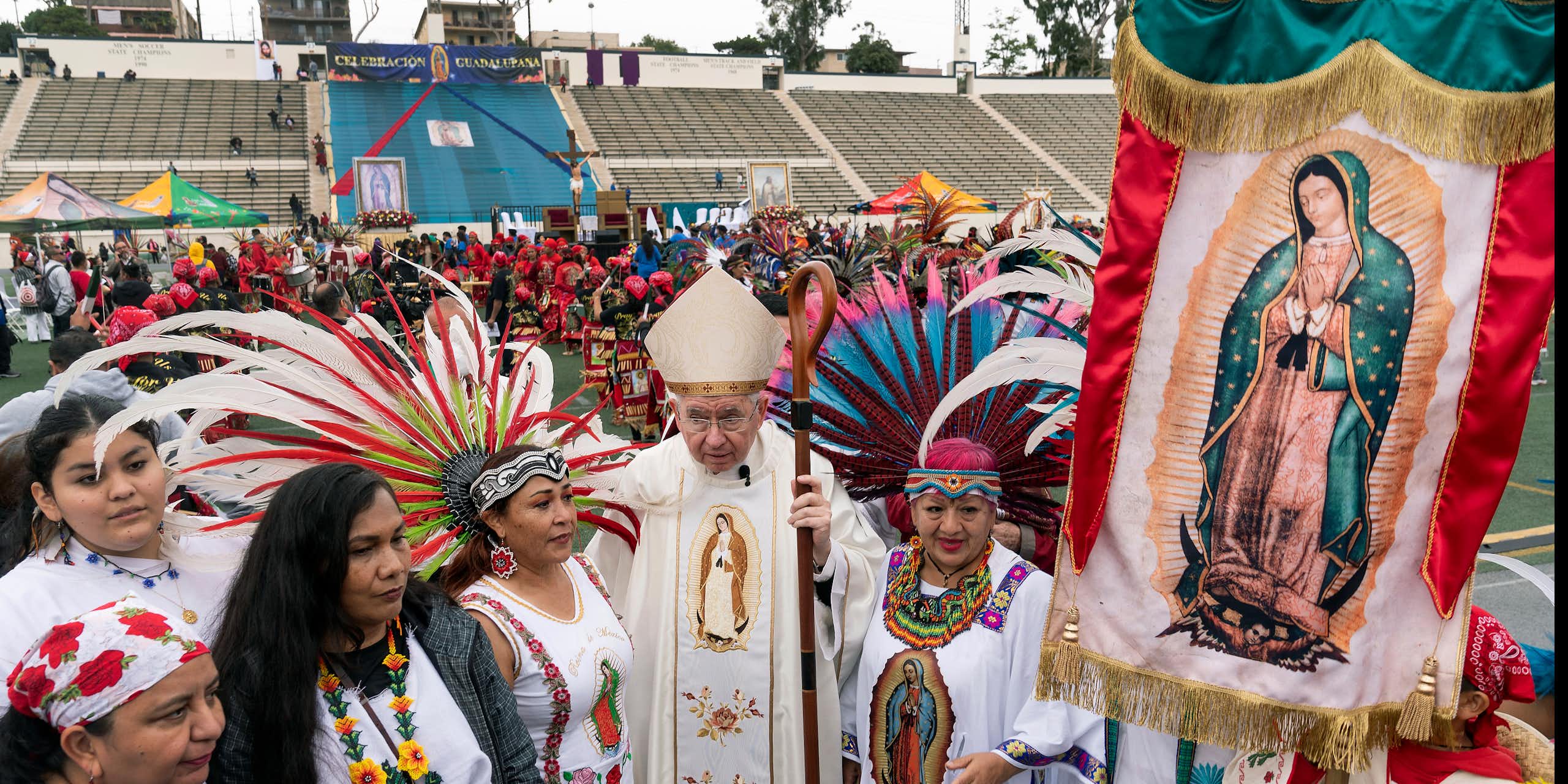 A man in white clerical vestments stands in an outdoor stadium, surrounded by women in white outfits holding a banner with a picture of a woman in a blue cloak.