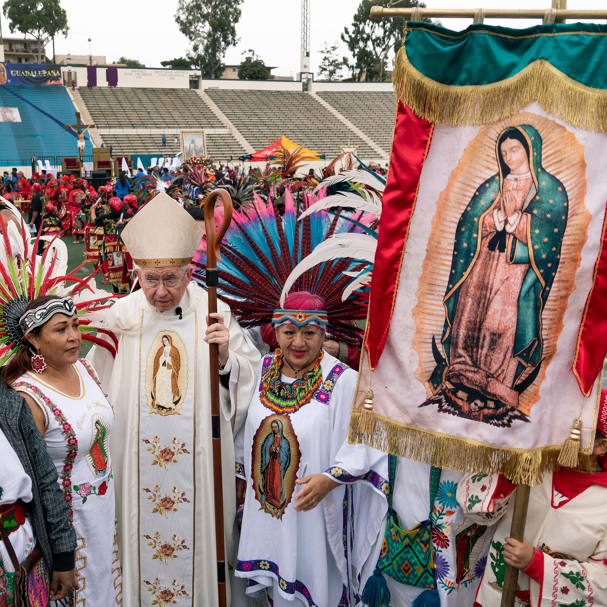 A man in white clerical vestments stands in an outdoor stadium, surrounded by women in white outfits holding a banner with a picture of a woman in a blue cloak.