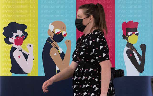 A woman in a face mask walks past a poster showing illustrations of masked people who have been vaccinated