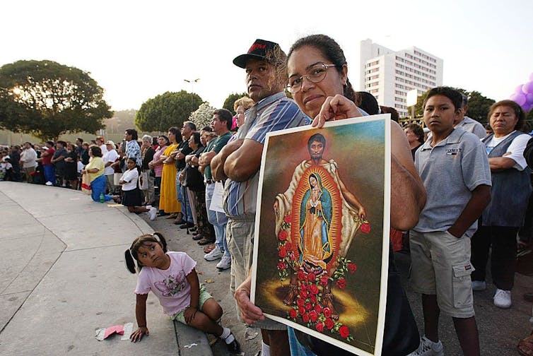 A woman holds a brightly colored poster of a man holding a picture of the Virgin Mary as she waits in a crowd in a concrete plaza.
