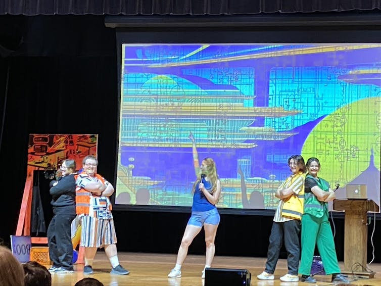 A group of people performing on a stage, wearing brightly colored costumes. The background is a screen projecting blue, green and yellow geometric shapes. The two performers on the left have their arms crossed and stand back to back, same on the right.
