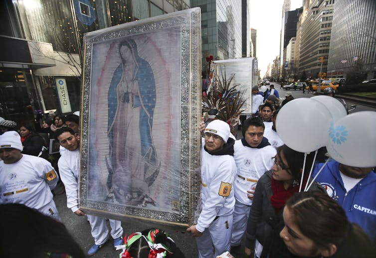 A handful of men in white attire walk down a city street carrying a banner with a faded picture of a woman in a blue cloak.