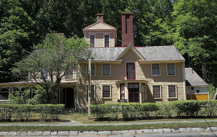 Large, old yellow house with big windows and a thick, tall chimney, flanked by trees.