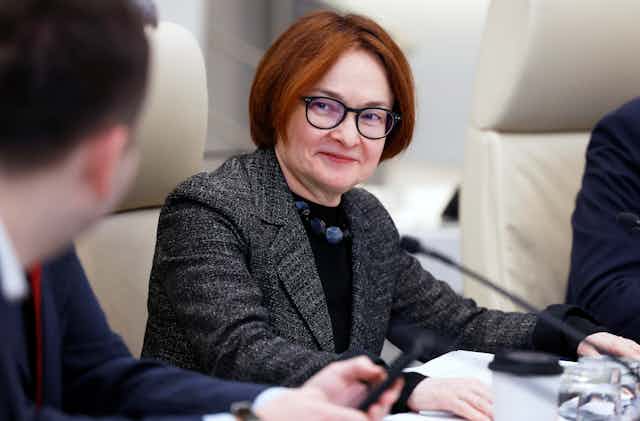 A woman with short auburn hair and glasses smiles while talking to a colleague.