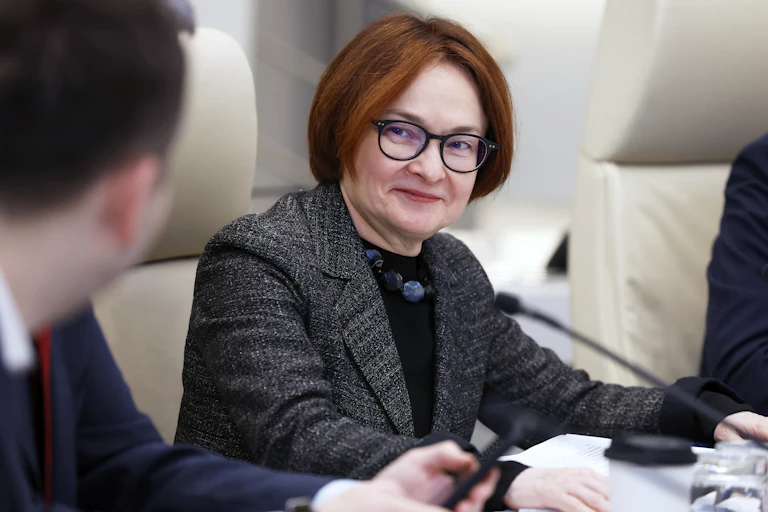 A woman with short auburn hair and glasses smiles while talking to a colleague.