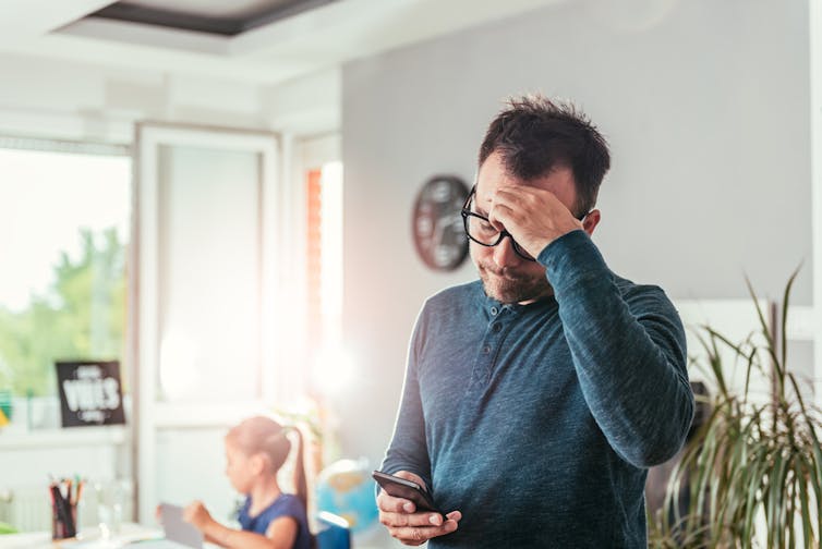 A worried father looks at his phone while his daughter sits in the background.