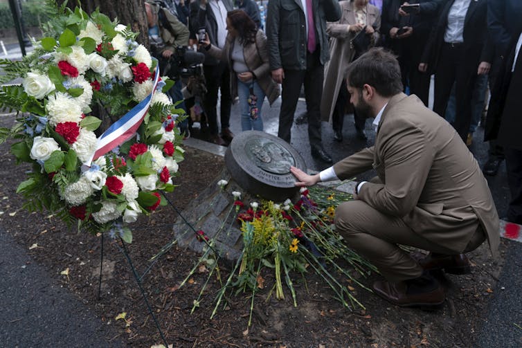 A man in a brown suit crouches down to touch a plaque strewn with flowers.