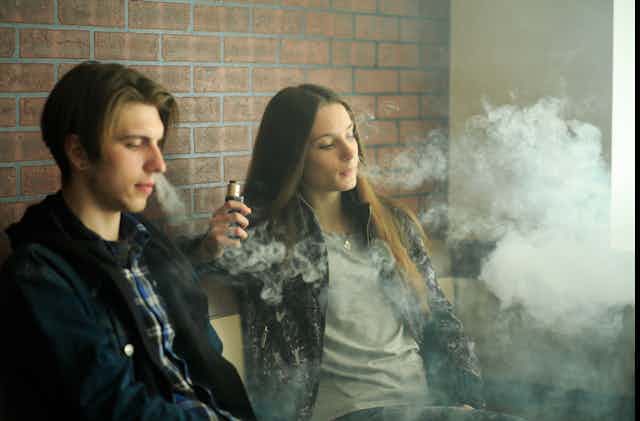 As puffs of smoke fill the air, two teenagers, one male and one female, smoke an e-cigarette while sitting against a tiled wall.