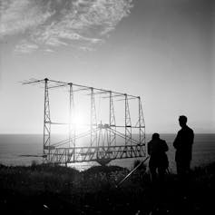 Black and white image of silhouettes of two people standing on a clifftop next to an antenna