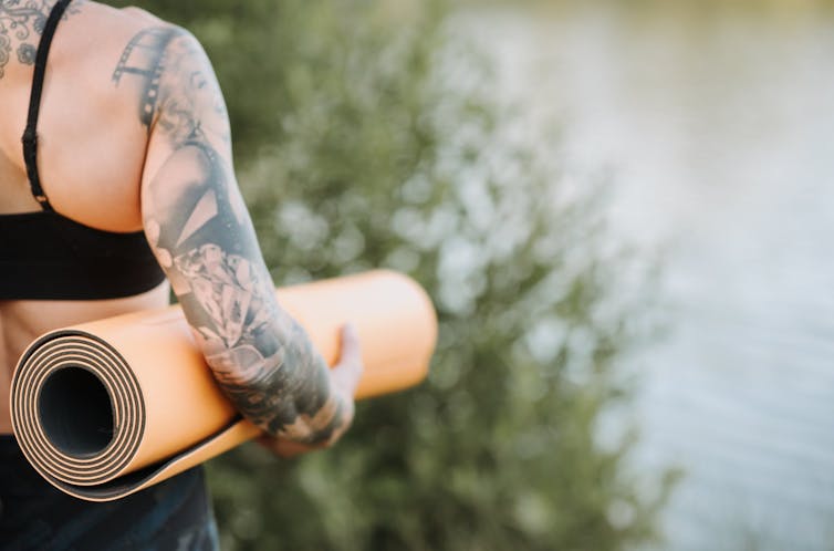 Woman with arm tattoo heading to sea, yoga mat under arm