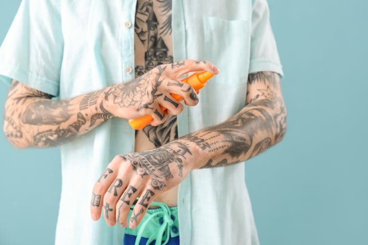 Tattooed man spraying sunscreen on arms.