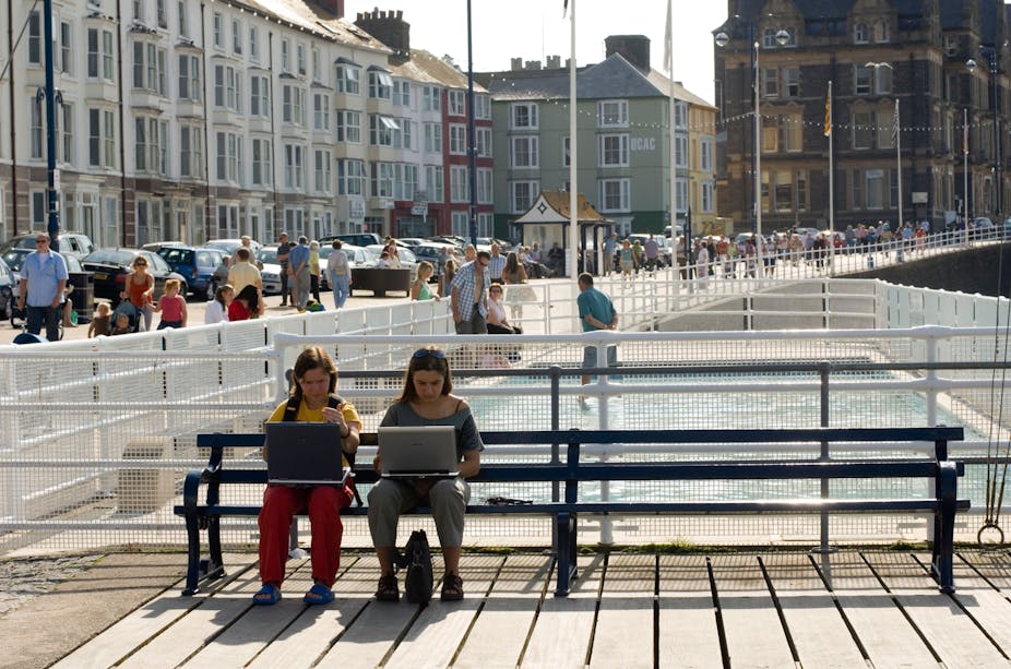Two women sit on a bench with laptops on their laps.