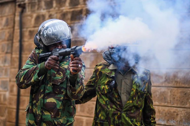 A man dressed in camouflage and a helmet written 'police' holding a firearm whose barrel is emitting a plume of smoke. A second man dressed similarly stands beside him