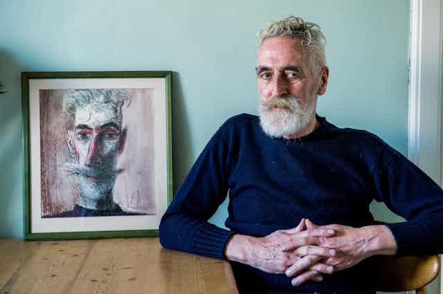 A striking grey-haired man – John Byrne – sitting at a table with a self-portrait 