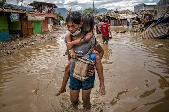 A woman carries a child on her back though flooded streets in a severely damaged neighborhood.