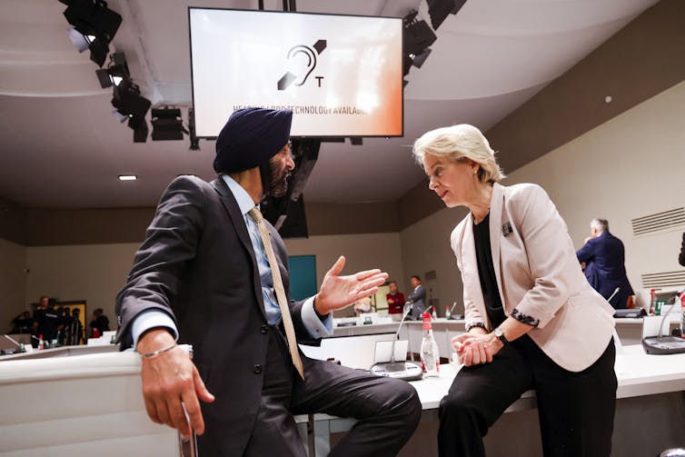 Banga, wearing a turban, and von der Leyen talk while sitting on the edge of a desk in a conference room.