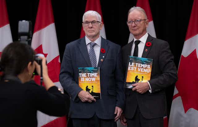 Two men in dark suits holding French and English versions of a report, standing in front of Canadian flags