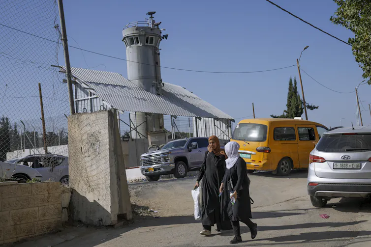 People walk past a fortified tower with cameras and weapons on top.