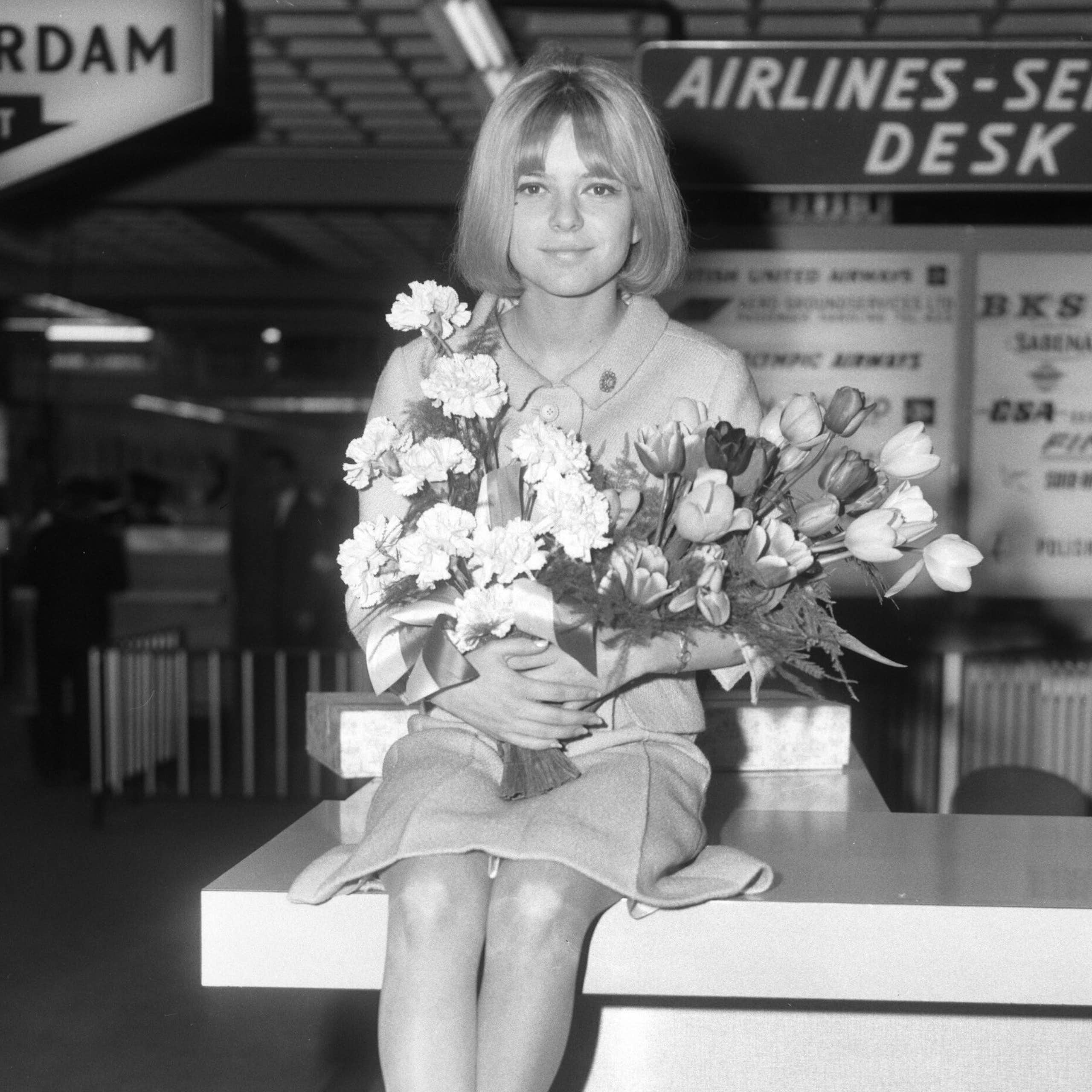 Una mujer joven sentada con traje chaqueta sobre un mostrador y un ramo de flores en las manos.
