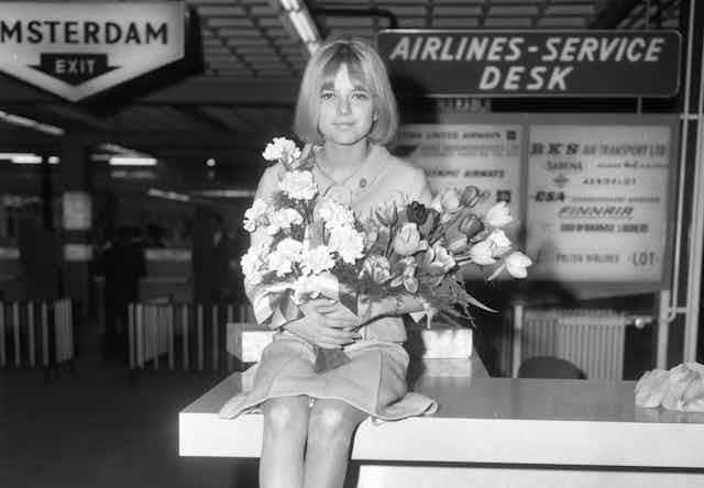 Una mujer joven sentada con traje chaqueta sobre un mostrador y un ramo de flores en las manos.
