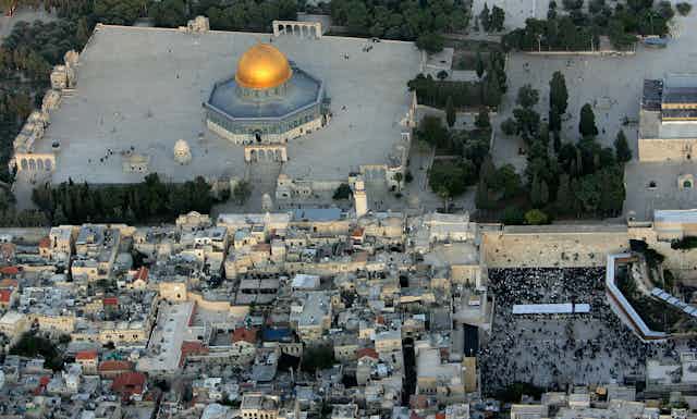 An aerial view shows many buildings, with a large golden dome on top of a building in a flared platform, adjacent to a limestone wall with many people crowding nearby it. 