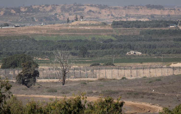 A fence divides rural, arid land, with trees and grass all around, and small mountains in the distance.