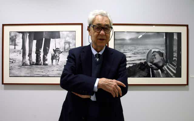 An old man with glasses (Elliott Erwitt) standing in front of two black and white photographs.