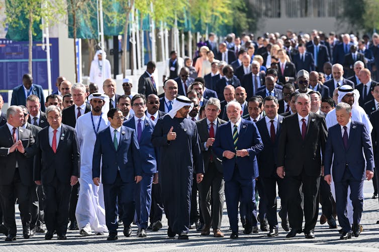 Several dozen older men in suits walk with al-Jaber, who is one of a few men in traditional Middle Eastern dress. There might be six women  in he photo, all, including Ursula von der Leyen, near the back.