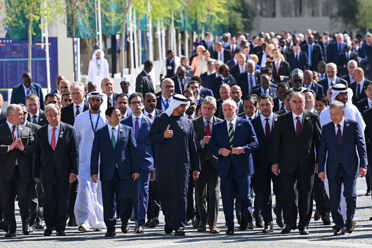 Several dozen older men in suits walk with al-Jaber, who is one of a few men in traditional Middle Eastern dress. There might be six women in he photo, all, including Ursula von der Leyen, near the back.