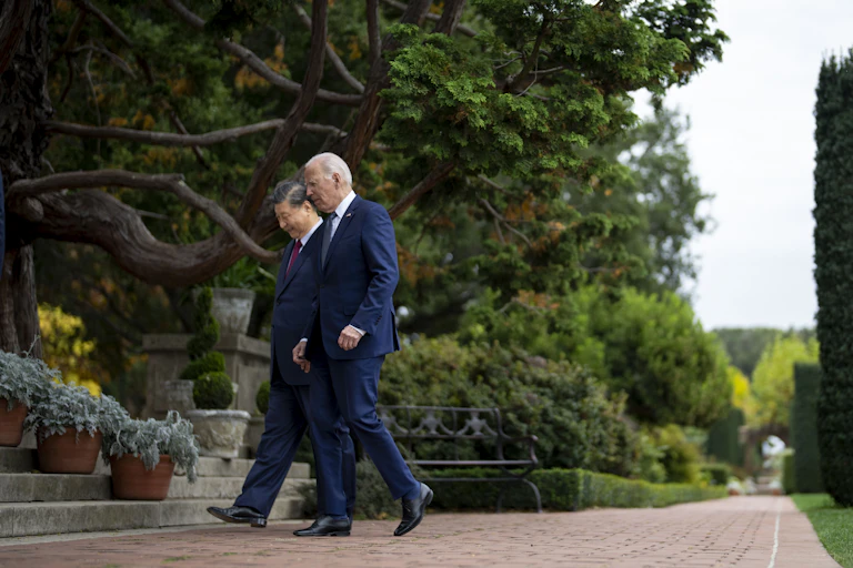 Two men seen walking side-by-side in front of a row of trees.