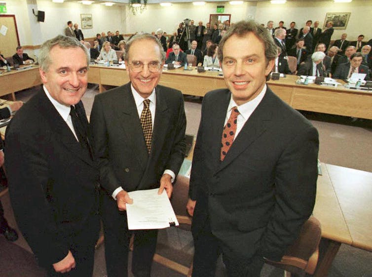 Three men wear dark suits and smile, together holding a white document and looking at the camera.