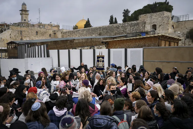A group of people, mostly women, seen standing near a wall and one is holding a rectangular object with two poles on either end high in the air.