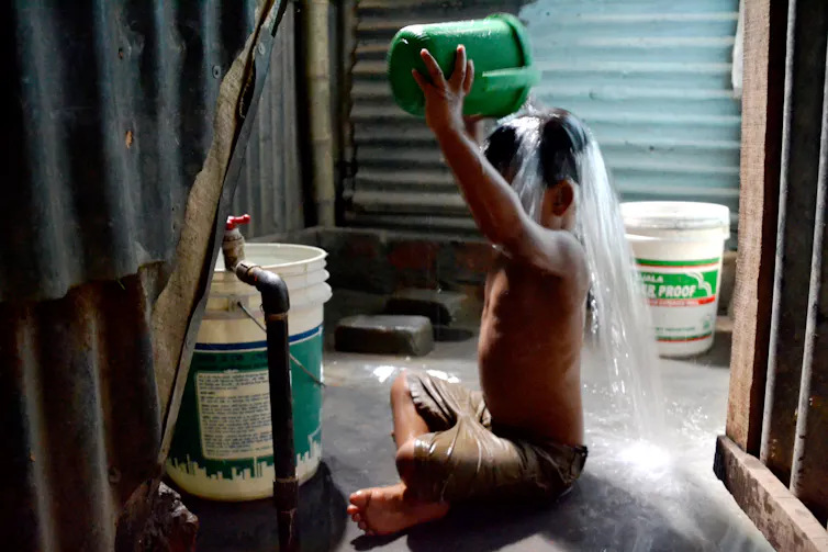 A boy sat surrounded by corrugated iron pouring a bucket of water over his head.