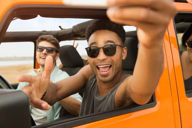 Young men on road trip, one holding arm out of window taking selfie