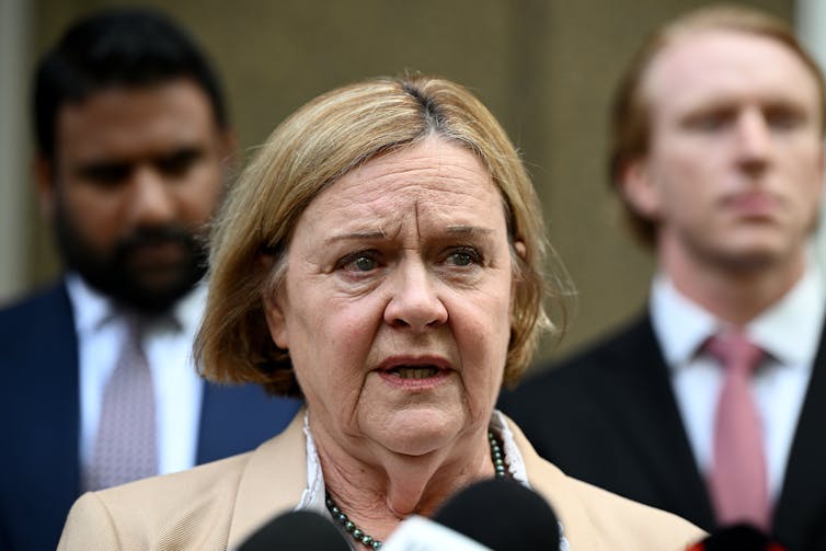 A woman with blonde hair addresses the media in front of two men in suits