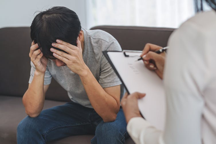 man sits with head in hands opposite clinician with clipboard checklist