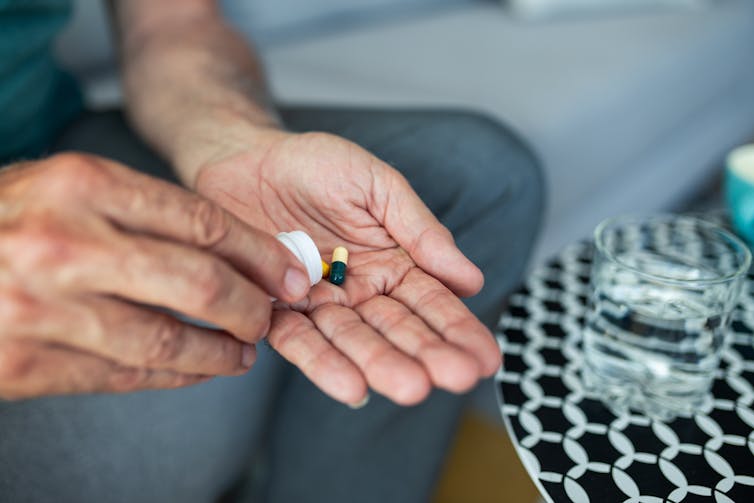 man's hands tipping out one capsule with glass of water nearby on table