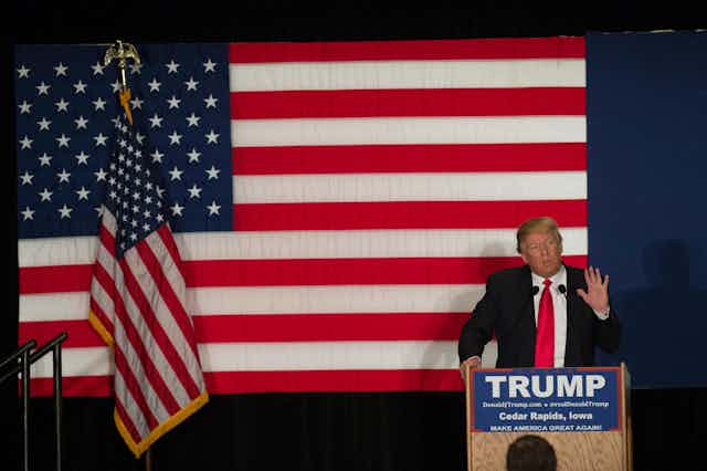Donald Trump in a suit, standing in front of a large US flag.