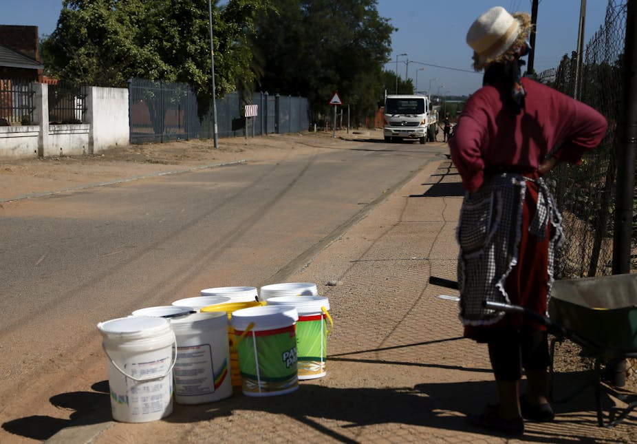 A woman stands on the side of the road with nine empty 20 litre buckets waiting for a water truck to come past so she can fill them