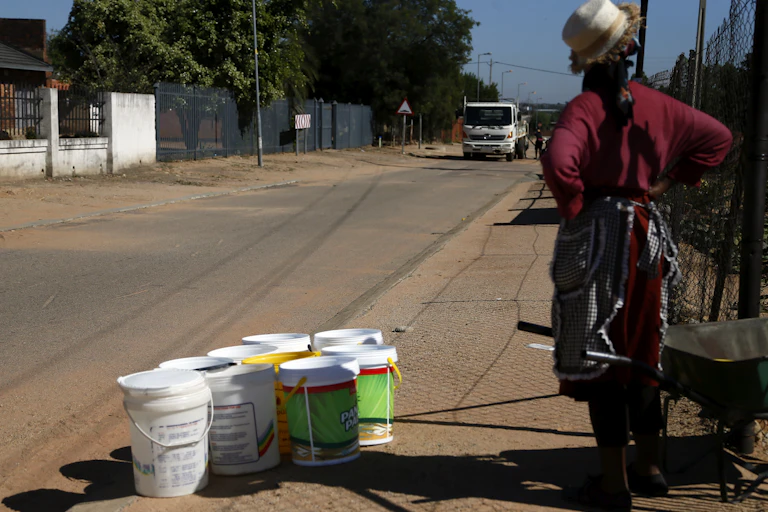 A woman stands on the side of the road with nine empty 20 litre buckets waiting for a water truck to come past so she can fill them