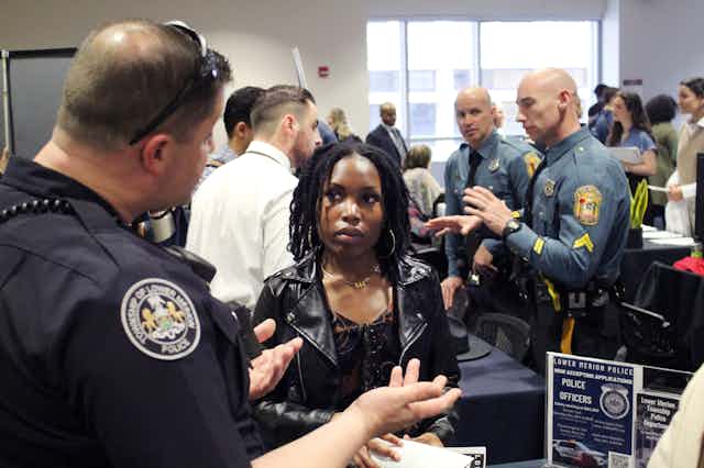 A young Black woman wearing a leather jacket listens as a uniformed officer talks to you