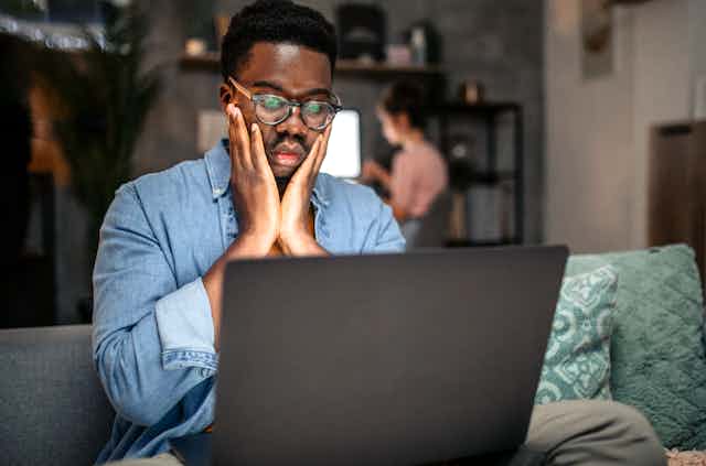 A man wearing glasses looks at a laptop with his hands on his cheeks call Sacha
