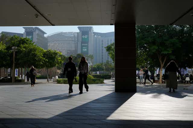 Two people in shadow seen walking towards the camera in front of a large building.
