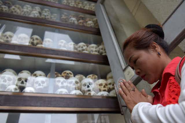 A Cambodian woman holds her hands together in prayer and looks straight ahead, standing in front of a four-tier glass cabinet that has human skulls stacked inside of it. 