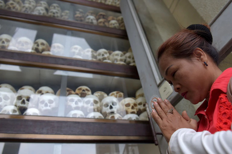 A Cambodian woman holds her hands together in prayer and looks straight ahead, standing in front of a four-tier glass cabinet that has human skulls stacked inside of it.