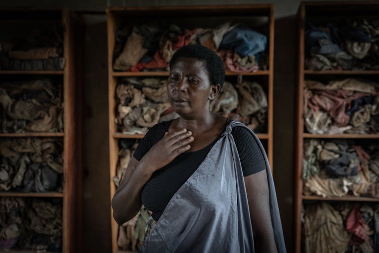 A black woman wearing a gray outfit that looks like a sari holds her hand to her chest and stands in front of shelves filled with old looking clothing.