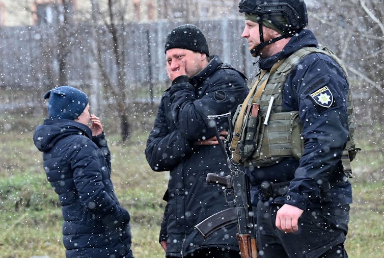 Three people wearing dark clothing, including one man with an army vest, stand in the snow. The woman and one man cover their mouths and look away, one man looks forward.