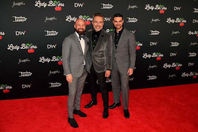 Three men in gray suits pose on red carpet.