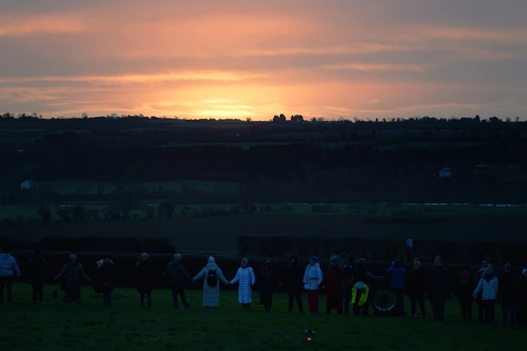 People holding hands and standing in a vast green field as the sun rises from afar.